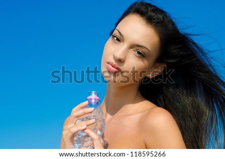 Beautiful brunette girl, with long hair in the wind, holding a bottle of water. Outdoors, blue sky background.