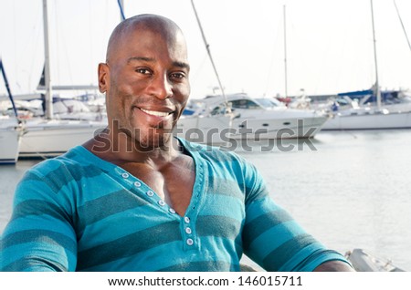 Summer marine scene with a handsome black man. Attractive man wearing a blouse with stripes at the sea side. Yachts in the background.