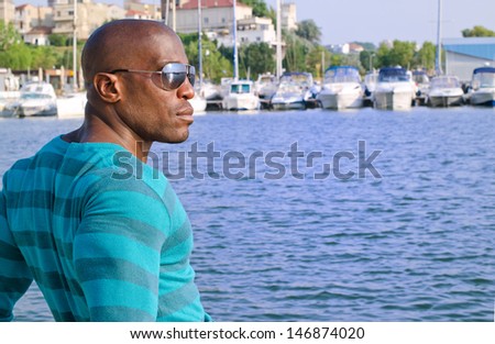 Summer marine scene with a handsome black man. Attractive man wearing a blouse with stripes at the sea side. Yachts in the background.