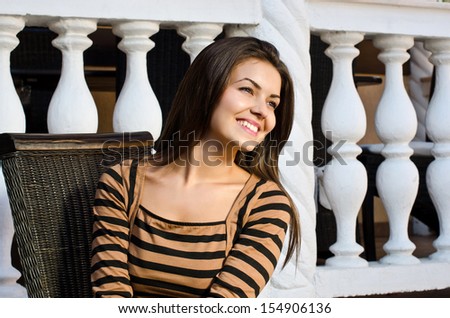 Girl smiling day dreaming on a chair. Beautiful woman sitting alone at a restaurant looking away. Outside, background nice architecture with columns.