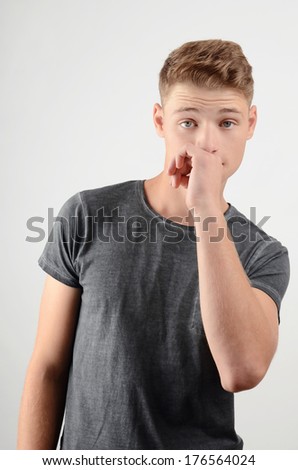 Young man smelling something bad. Disgusting odor. Guy frowning and holding his hand at his nose. Man in grey t-shirt with nasty face expression.