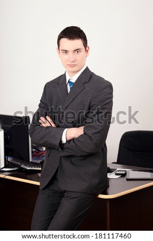 Young business man with arms crossed. Man in suit standing in front of his desk confident.
