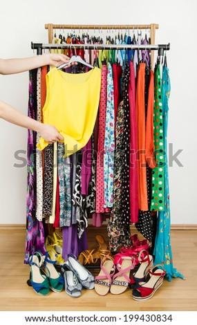 Woman hand picking up a yellow blouse to wear. Summer dresses and sandals in a wardrobe. Dressing closet with colorful clothes and shoes nicely arranged on a rack.
