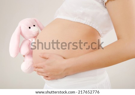 Close up on pregnant belly with toy. Woman expecting a baby with a cute pink rabbit toy peaking at her belly. Easter baby.