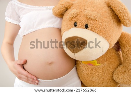 Close up on pregnant belly and a big teddy bear. Woman expecting a baby with a cute big teddy bear hugging her belly.