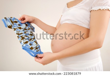 Close up on pregnant belly with baby clothes. Woman expecting a baby dressed in white holding a cute camouflage baby onesie.