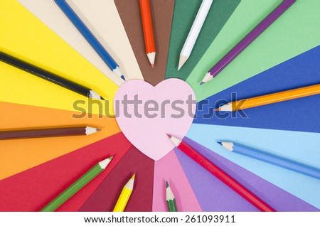 Color pencils on paper with pink heart memo note. All colors pencils arranged in a circle on rainbow color paper with blank post it note.