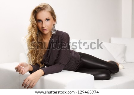 beautiful blond woman lying on a white couch, she is dressed in leggings, shirt and shoes in the heel, interior photos, fashion photos