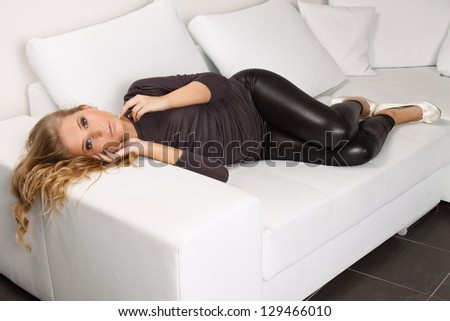 beautiful blond woman lying on a white couch, she is dressed in leggings, shirt and shoes in the heel, interior photos, fashion photos