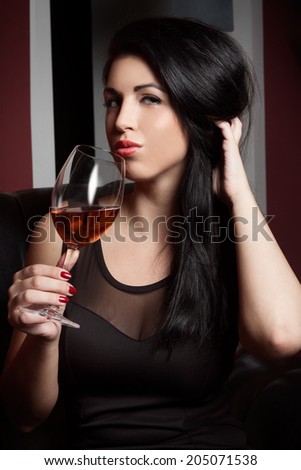 Beautiful brunette in a black dress, with glass of wine sits on a armchair, in a interior, wine bar, coffee house, fashion photography