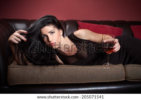 Beautiful brunette in a black dress, with glass of wine lies on a couch, in a interior, wine bar, coffee house, fashion photography