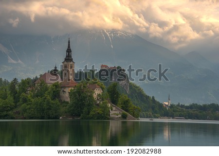 View to Lake Bled\'s church and castle on a dramatic sunrise color falling over the background mountains.
