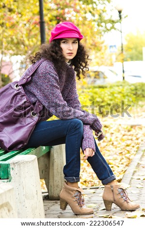 Young stylish woman dressed in oversized knitted sweater, skinny jeans, peaked cap and heels with big bag sitting on bench in city park. Fashion beautiful girl