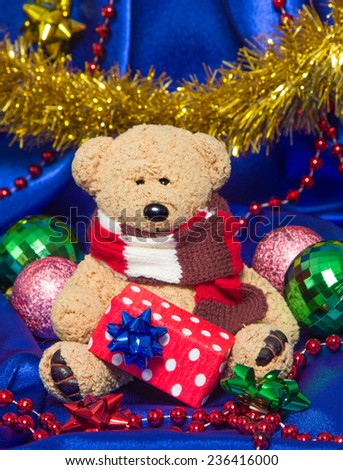 Charming small teddy bear holding gift box in paws surrounded by Christmas balls, tinsel and beads