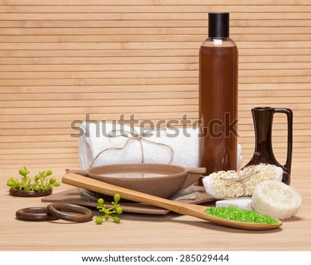Spa and pampering products and accessories: sea salt, pumice, loofah, wisp of bast, bamboo utensils with water, crock, shower gel, bath towel on wooden surface