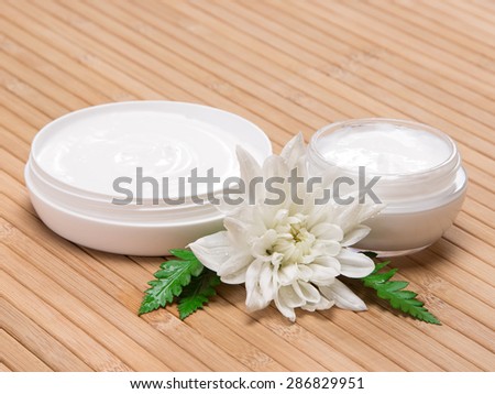 Natural moisturizing skin care products. Closeup of two open jars filled with cream next to wet white flower and fern leaves on wooden surface