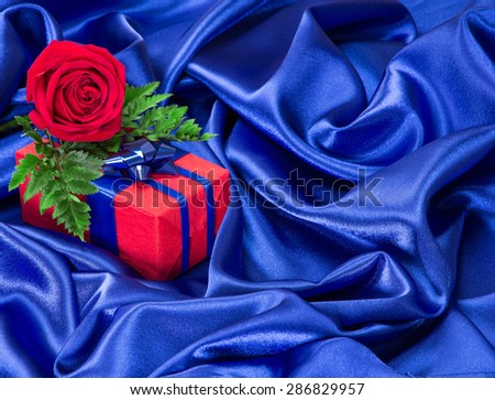 Red gift box with blue bow and rose surrounded by dark blue silk fabric. Copy space