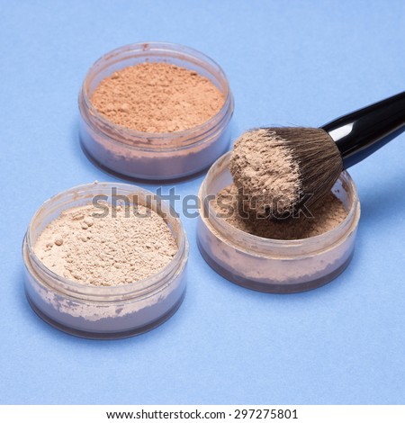Close-up of makeup brush and jars filled with loose cosmetic powder different shades on blue background