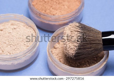 Close-up of makeup brush and jars filled with loose cosmetic powder different shades. Blue background. Side view, focus on bristle brush, shallow depth of field