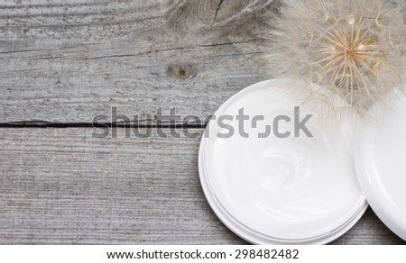 Concept of light like a dandelion skin care cream. Open jar filled with cream and big fluffy dandelion on old wooden planks. Fresh light texture of cosmetic product for gentle skincare. Copy space