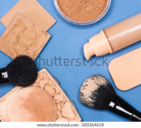 Close-up of concealer, corrector, open cream foundation bottle and jar filled with loose powder, crushed compact powder, makeup brushes and cosmetic sponge on blue surface