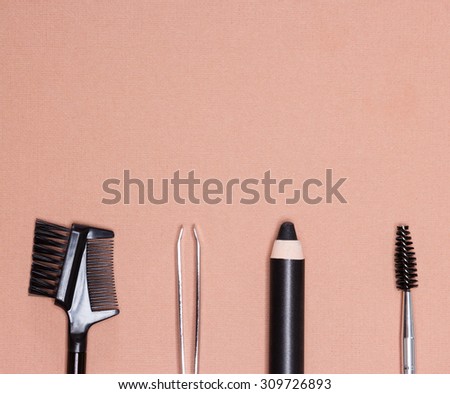 Accessories for care of brows: eyebrow pencil, tweezers, brush and comb on peach colored textured surface. Eyebrow grooming tools. Copy space in the upper portion of the image