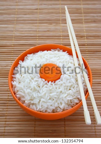 Orange bowl of cooked rice decorated with carrots and sticks on bamboo napkin.