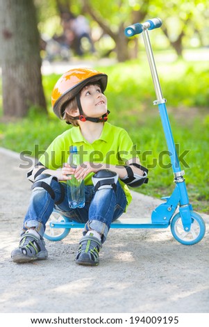 Positive small boy sits on scooter and looks up on path in city park