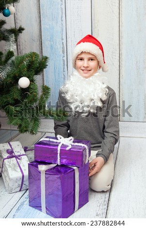 The boy in white beard and red cup sits at New Year tree with his presents.
