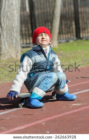 Small boy sits on race track of school stadium with skateboard in spring time