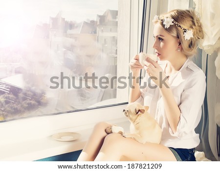 beautiful girl with small dog on knees with a cup of coffee looking through window