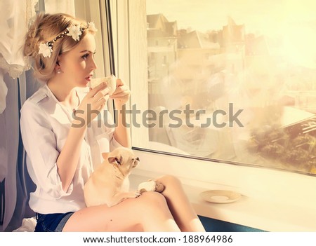 beautiful girl with small dog on knees with a cup of coffee looking through window