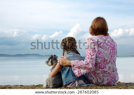 Mom and daughter sit embracing on the lake and look at the water. Near them sits their small dog.