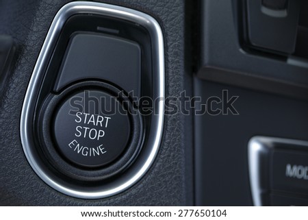 A close shot of a modern car engine start and stop button. The button is round and black with start and stop written in black. It is placed upon a black console and features clean lines.
