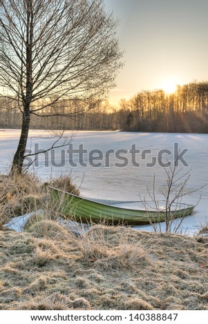A boat filled with ice at the edge of a snow covered and frozen over lake. It is early spring in Sweden. The sun rises behind a small hill casting light upon the lake.
