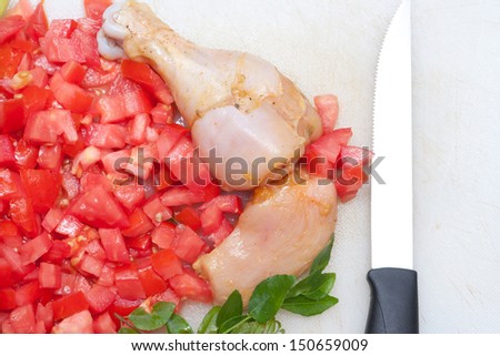 Raw chicken on cutting board with tomatoes onions and curry leaves. vegetables and herbs. Knife on cutting board.