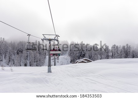 Ski lift with passengers in the chair traversing a snow covered mountain slope as it starts the ascent over the evergreen pine plantations to the distant summit and start of the ski runs