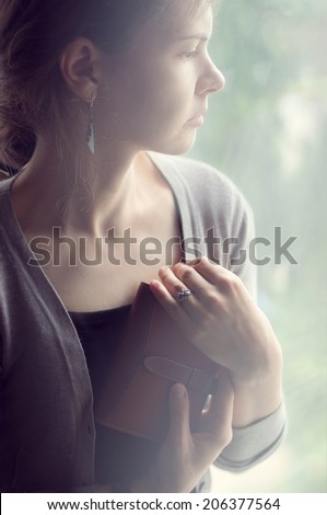 background girl sitting and looking out the window and holding a notebook, clutched to her chest, closeup body and arms, flare