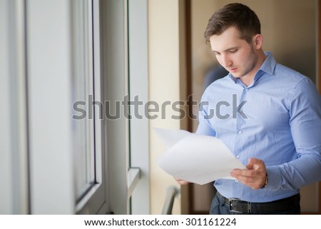 Smiling businessman standing by the window in office and reading paper