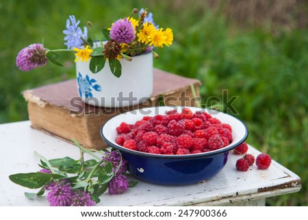 Blue bowl with raspberries, old book and cup with field flowers on the rustic white chair with the haymow on the background .