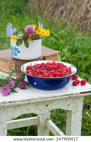 Blue bowl with raspberries, old book and cup with field flowers on the rustic white chair with the haymow on the background .
