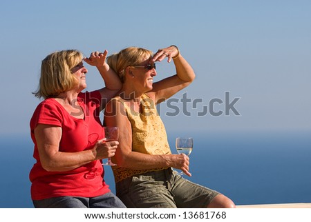 Two Senior Ladies Looking Off Camera with a Glass of Wine, against a Blue Sky and Sea Background