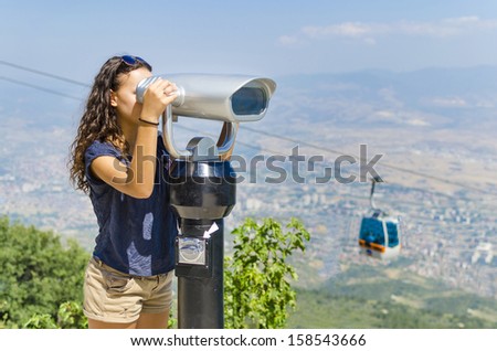Great Spectators - Stock Image. Beautiful girl  is looking at coin operated binocular at the top of Vodno mountain, Skopje, Macedonia with cable car at background