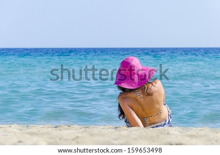 Attractive, fit and tanned girl sitting on white sandy beach in the sun adjusting pink straw hat on vacation. Blue sky, blue sea in Spain.