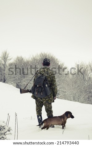 Male hunter in camouflage clothes walking on the snow field with hunting rifle during a hunt, dog follows him, foggy weather