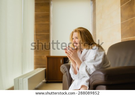 Beautiful young blonde girl in a white robe looking out the window, and drink of that Cup. laughs.