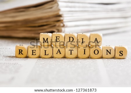 Concept of dices with letters forming words: Media Relations. Generic newspaper background with some blurred text on the bottom and paper stack in the back.