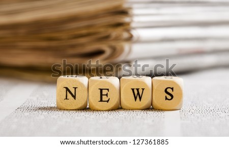 Concept of dices with letters forming word: News. Generic newspaper background with some blurred text on the bottom and paper stack in the back. Dices made from wood with natural imperfections.
