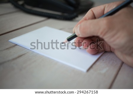 Male hand with pencil writing on blank paper note pad, on wooden table. Selective focus with shallow depth of field.