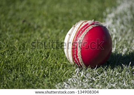 An old red and white cricket ball with worn stitching sits abandoned on the sports field boundary